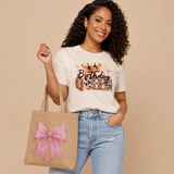 A woman wearing a white graphic t-shirt that reads 'Birthday Queen' with animal print patterns, paired with light blue jeans and holding a beige tote bag with a pink bow, against a solid beige background.