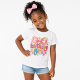 A young girl with curly hair and a pink bow is wearing a white graphic t-shirt that says 'birthday girl' in colorful bubble letters with a bow design, paired with denim shorts, against a plain white background.